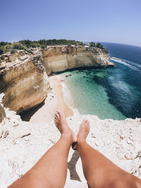 Low section of person relaxing on cliff by sea against clear blue sky