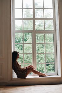 Side view of woman sitting on window sill at home