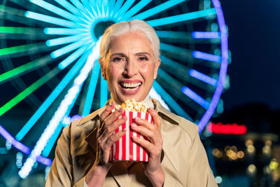 Portrait of smiling woman holding popcorn against ferris wheel