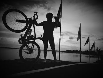 Man standing with bicycle by flag on roadside against lake