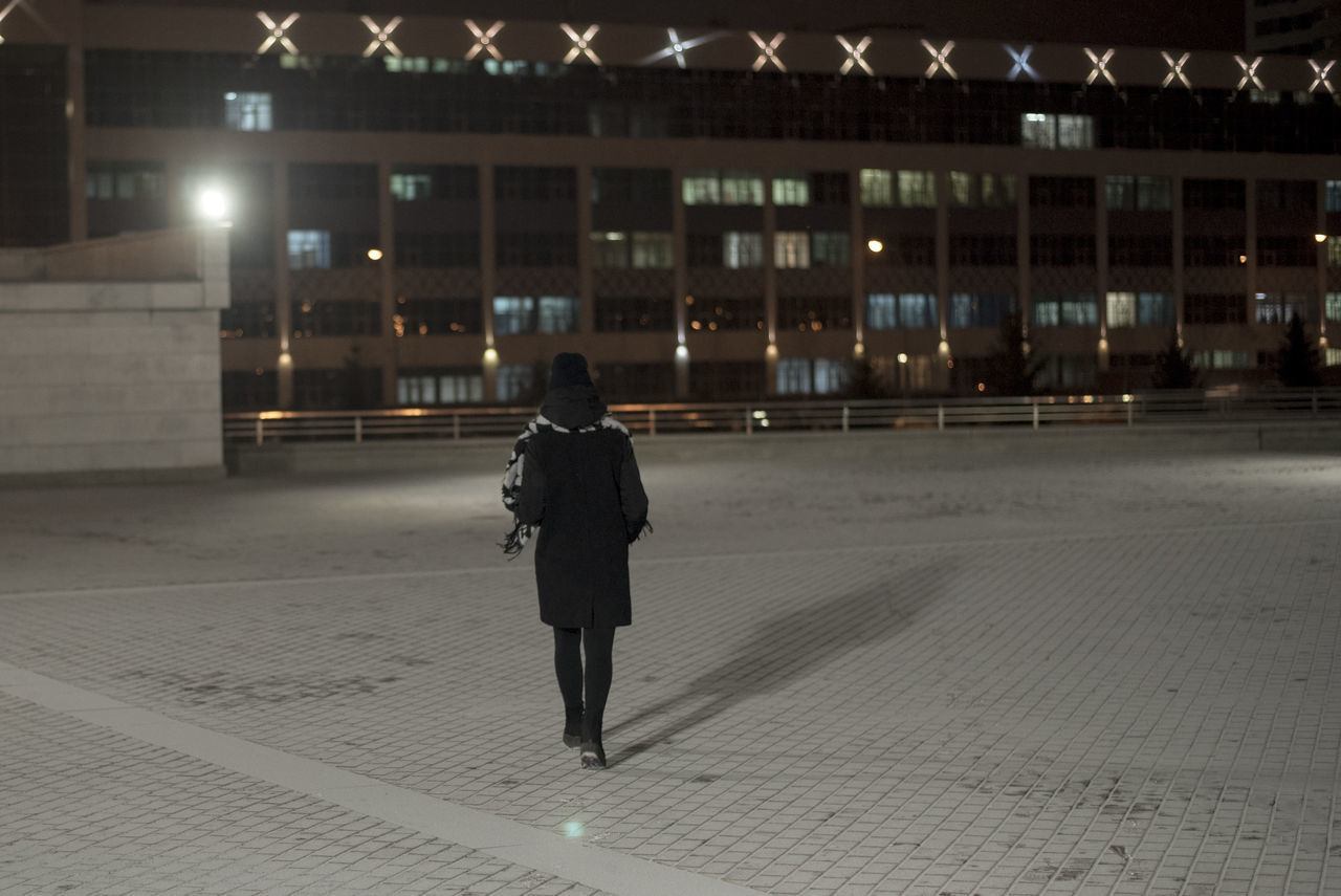 REAR VIEW OF WOMAN WALKING ON STREET AGAINST ILLUMINATED BUILDINGS
