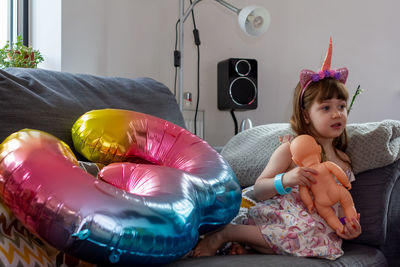 A cute baby girl sitting on a sofa with a colourful balloon and a doll barefoot