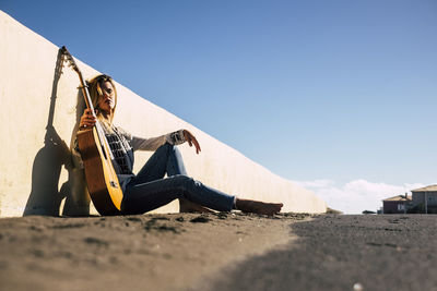 Woman sitting with guitar while leaning on wall against clear sky on sunny day