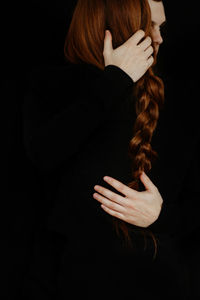 Back view of unrecognizable crop tender boyfriend tenderly embracing redhead girlfriend while standing in dark studio on black background