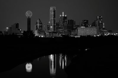 Illuminated city buildings by river against clear sky at night