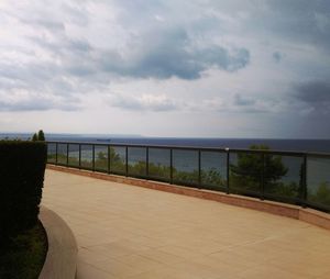 Empty pathway along calm sea against cloudy sky