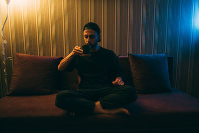 A young man drinks tea sitting on a sofa