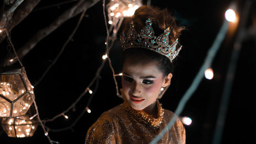 Close-up of beautiful model wearing crown amidst illuminated string lights at night