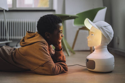 Cheerful boy in front of illuminated ai robot while lying on floor in innovation lab