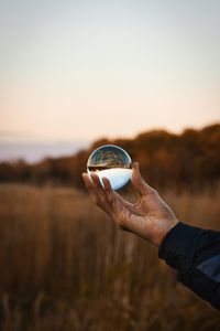 Close-up of hand holding crystal ball on field against sky during sunset