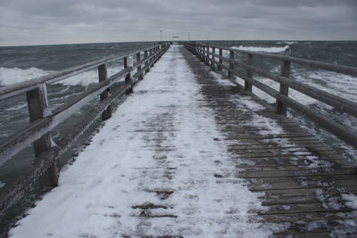 Snow covered pier over sea against cloudy sky