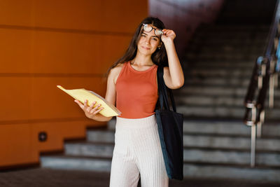 A beautiful brunette with a folder in her hand stands near the stairs at a university or college