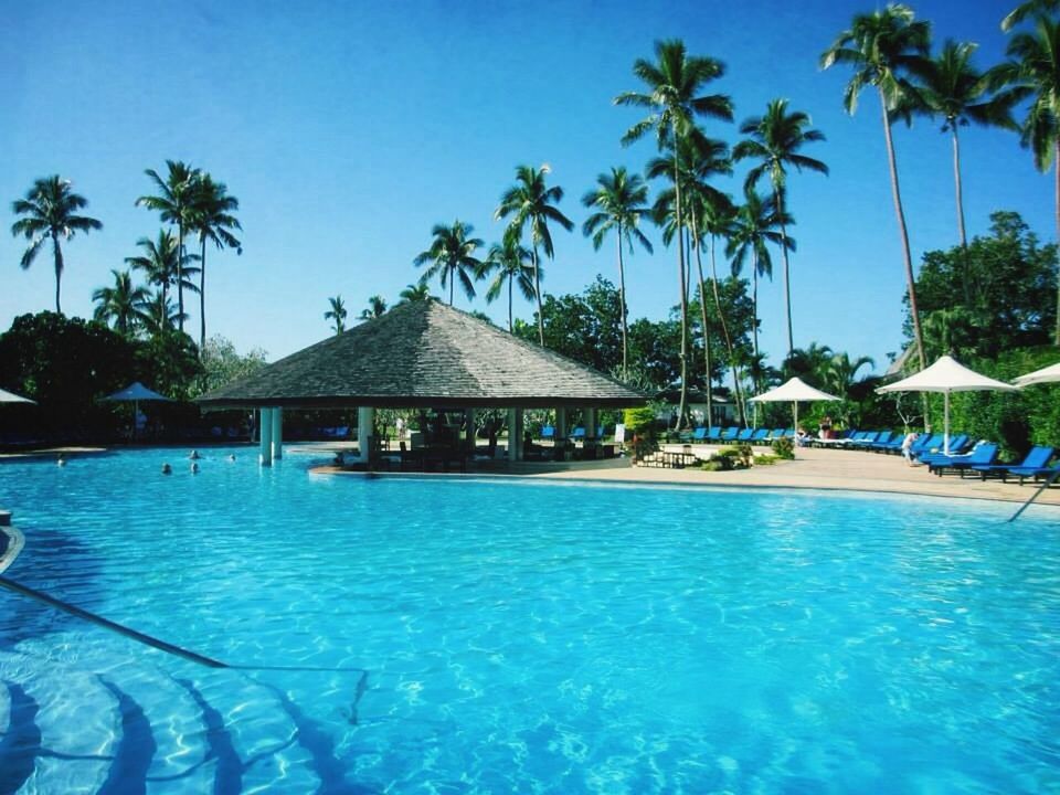 water, tree, palm tree, blue, swimming pool, sea, building exterior, built structure, tourist resort, architecture, sunlight, clear sky, beach, turquoise colored, waterfront, vacations, nature, tranquil scene, incidental people, house