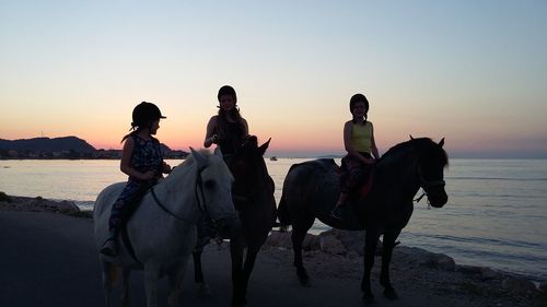 Sisters riding horses at beach against clear sky during sunset