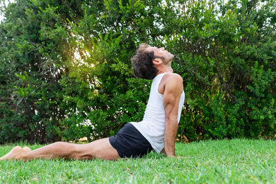 Side view of middle aged man in tank top and shorts stretching back in urdhva mukha svanasana pose on grassy lawn near green bushes during yoga session on summer day in park