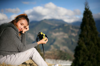 Portrait of young woman sitting on rock with camera against sky
