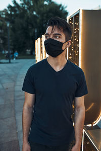 Young man standing in the city center in the evening looking away wearing the face mask
