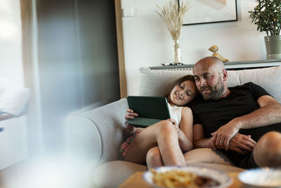 Smiling daughter sharing digital tablet with father while sitting on sofa in living room