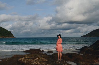 Rear view of woman standing on shore at beach against cloudy sky