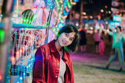 Portrait of smiling young woman against illuminated multi colored lighting equipment
