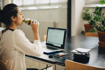 Businesswoman drinking juice while using laptop at desk in office