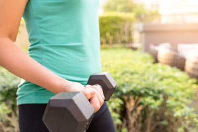 Midsection of woman holding dumbbell while exercising in backyard