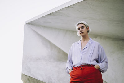 Portrait of non-binary person standing with hands in pockets against wall