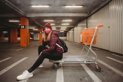 Young woman with mask in urban outfit sitting on shopping cart