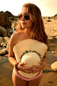 Young woman wearing sunglasses holding hat while standing at beach