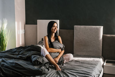 Young caucasian brunette with long hair in pajamas is sitting on the bed in the morning and smiling