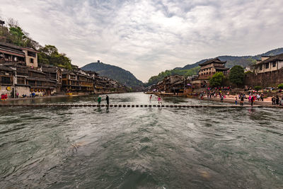 People at riverbank against cloudy sky