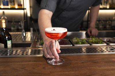Professional bartender offers a glass of delicious alcohol cocktail with foam and flower petal.