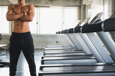 Midsection of shirtless man standing by treadmills in gym