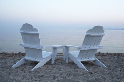 White adirondack chairs on sand at beach against sky during sunset
