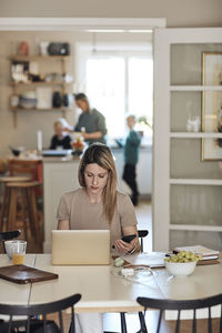 Businesswoman holding smart phone while working on laptop at home