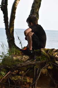 Boy sitting on rock by sea against sky