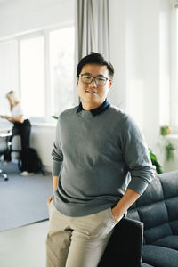 Portrait of smiling young businessman with hands in pockets leaning on sofa at office