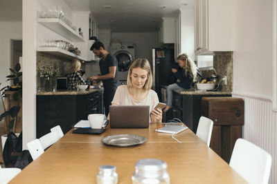 Mid adult woman holding mobile phone while using laptop at table with family in kitchen at home