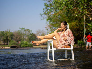 Woman sitting on a tree by plants against sky