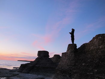 Low angle view of silhouette mature woman standing on rock at beach against sky during sunset