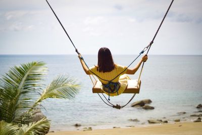 Rear view of woman on swing at beach against sky