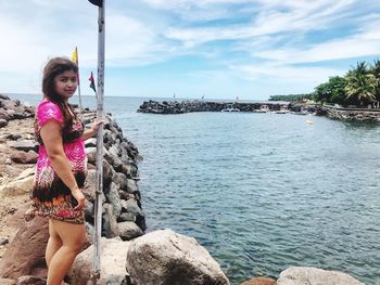 Portrait of young woman standing at rocky shore against sky