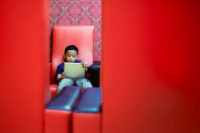 Boy sitting with digital tablet on chair against wall