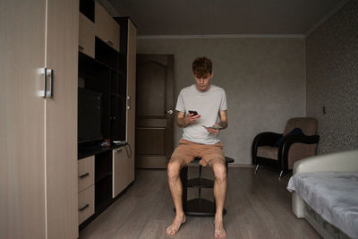 A guy at home in pajamas sits in the middle of the room with a phone and money in his hands