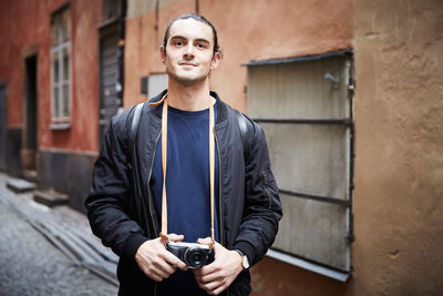 Portrait of young man holding camera while standing by building in alley