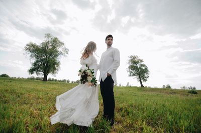 Side view of wedding couple standing on grassy field against sky