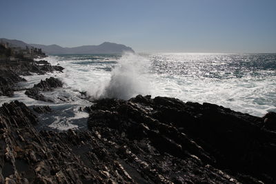 Waves splashing on rocks at shore against clear sky