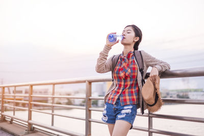 Young woman drinking water while standing against railing and sky