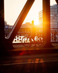 Woman walking with bicycle on bridge in city during sunset