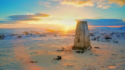 Wooden post on snow covered land against sky during sunset